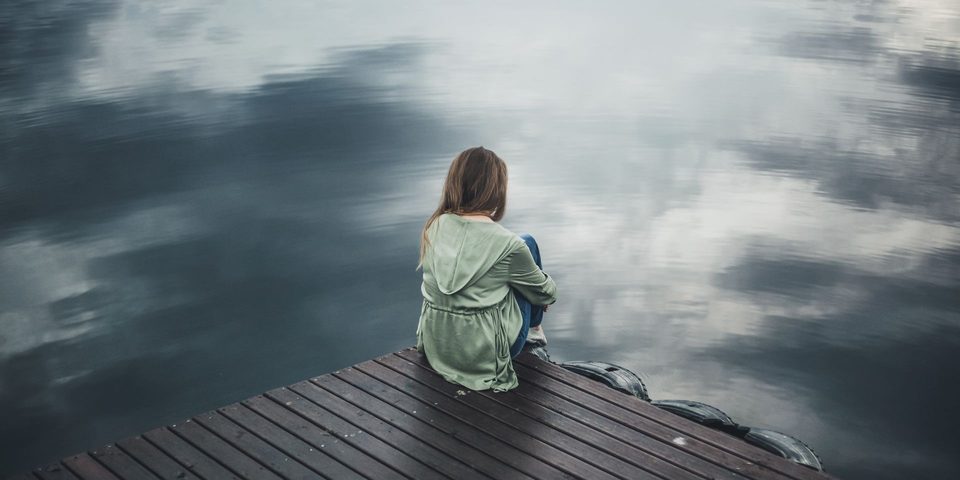 A woman sitting alone on a wooden dock, looking out at a calm lake